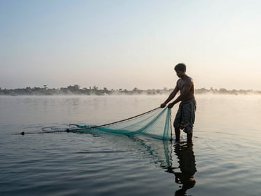 Environmental wide shot of a fisherman at dawn, hands pulling a net from still haor water, soft morning light, mist above the surface, natural teal tones