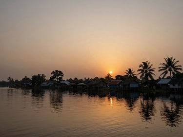 Dusk over a haor village, silhouettes of homes and palms reflected in still water, warm amber light fading at the horizon, quiet and wide