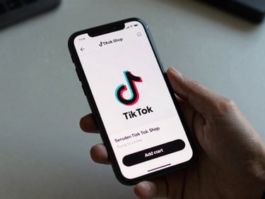 Mobile phone screen displaying a TikTok Shop product listing page with add-to-cart button visible, hand holding the phone against a neutral desk surface, cool daylight, tight crop
