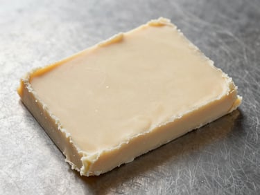 Macro overhead of solidified cocoa butter slab on a clean steel surface under a north-facing diffused studio light — pale ivory color, crystalline surface texture visible, no styling