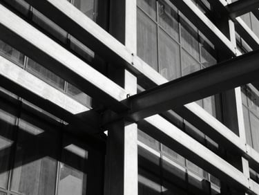 Abstract architectural crop of steel beams meeting a glass facade, sharp geometric lines, direct hard sunlight, high-contrast monochrome.