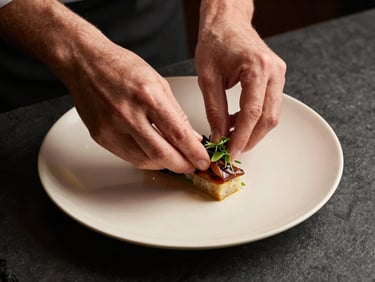 Close-up overhead shot of a chef's hands carefully placing garnish on a plated dish set on a dark slate surface, soft warm tungsten light from the left, precise and deliberate movement, cream-toned porcelain plate