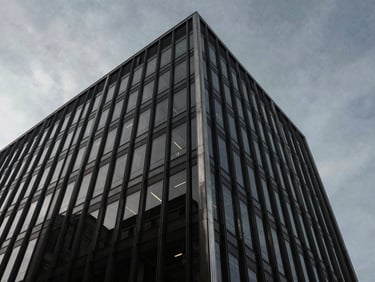 Dramatic low-angle shot of a dark glass office tower facade reflecting a quiet gray sky, premium corporate aesthetic