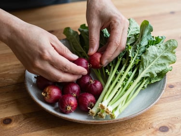 Overhead shot of hands styling a rustic plate of fresh seasonal vegetables on a wooden table, soft north-facing daylight, rich crimson and forest green tones, shallow depth of field.