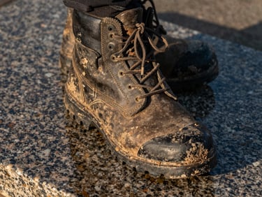 Close-up of a weathered boot sole on wet granite slab, laces mud-caked, late afternoon harsh sidelight casting deep shadows across the tread pattern, gear in hard use, no person visible above the ankle
