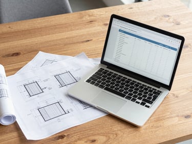 Clean overhead table shot of a working session with technical blueprints, a laptop displaying a cap table spreadsheet, and warm wood tones, bright natural daylight, 35mm.