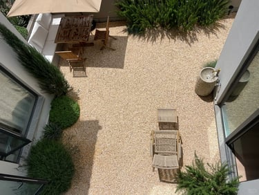Minimal outdoor seating area with gravel ground, wooden furniture, umbrella shade and green shrubs f