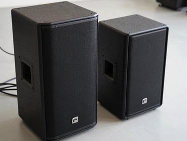 Close-up overhead shot of a professional PA speaker and subwoofer pair on a neutral light-grey floor, cables visible, bright studio lighting, no people