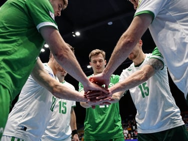 Intense team huddle of handball players in green and white uniforms, hands stacked in the center, dramatic sports hall lighting, high-contrast