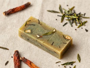 Overhead flat lay of a Matcha & Manjistha Ritual Bar, sliced to show botanical inclusions, on a raw linen cloth with scattered green tea leaves and Manjistha root pieces, soft sun-drenched natural light, macro texture, shallow depth of field