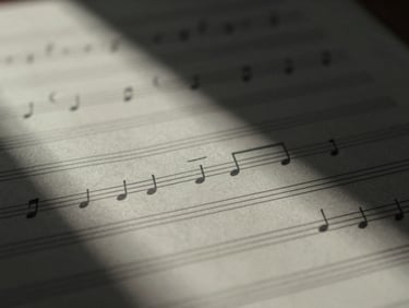 Macro close-up of manuscript paper with handwritten musical notes under soft natural window light, deep shadows, desaturated monochrome tones.