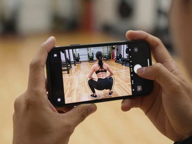Close-up of hands holding a smartphone displaying a video of a squat execution with overlay drawing lines for posture correction, warm gym background.
