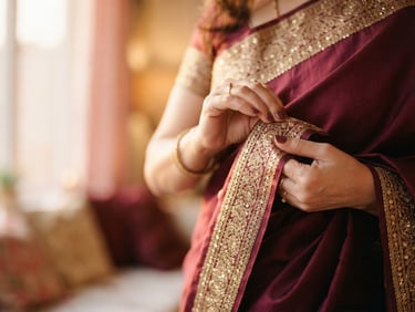 Elegant close-up of hands gently arranging the gold-threaded border of a rich maroon silk saree, soft golden-hour light, shallow depth of field.