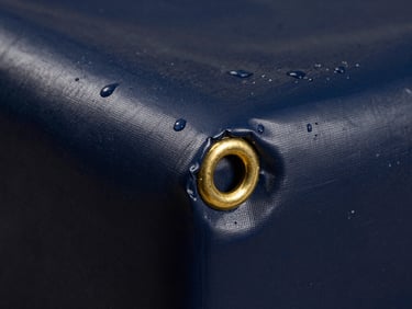 Macro shot of heavy-duty dark blue waterproof tarpaulin material, reinforced brass grommet in the corner, water droplets beaded on surface