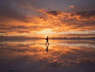 Cinematic wide shot of the Uyuni salt flats at sunset, perfectly reflecting the deep orange sky and a lone traveler walking.
