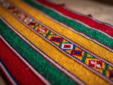 Macro shot of traditional Bolivian aguayo textile, focusing on the intricate red, yellow, and green hand-woven patterns under soft, warm side light.
