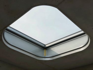 Tight framing of a custom frameless glass skylight curb edge, structural silicone joint visible between glass and steel frame, daylight flooding through from above, no people, concrete ceiling surround