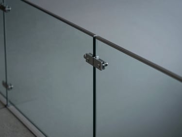 Tight architectural shot of a frameless glass railing post connection at a stair landing, cool even studio light, concrete floor visible below the glass panel, structural clip hardware in sharp focus, no people