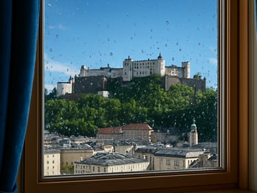 Crystal-clear window pane looking out to the Salzburg fortress, water droplets bead on the glass, bright daylight, 35mm