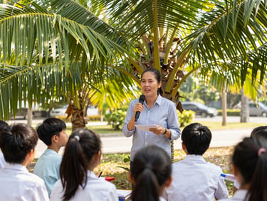 A teacher leading a lesson outdoors with students under a palm tree in Thailand, warm tropical sunlight.