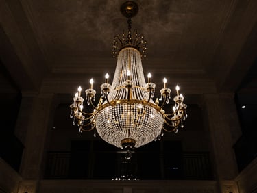 Low-angle shot looking up at a monumental brass and crystal chandelier glowing warmly in a grand hotel lobby, deep shadows, high contrast, low horizon.