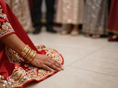 Photographer's perspective at floor level, close focus on a bride's hands resting on embroidered red fabric, gold bangles catching warm ambient light, soft bokeh background of gathered family