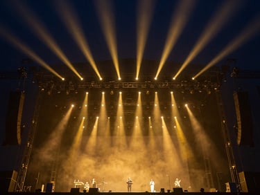 Wide-angle view of massive illuminated concert stage rigging, deep navy backlighting, golden spotlights cutting through atmospheric haze, extreme scale