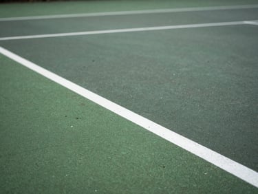 Close-up of a weathered green macadam tennis court surface, white painted baseline, soft natural overcast UK daylight.