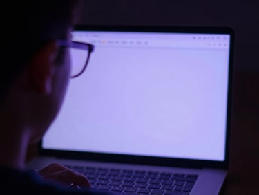 Macro over-the-shoulder view of a student studying on a laptop, soft violet screen glow reflecting on their glasses, dark ambient background.