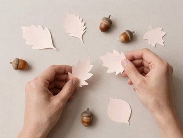 Overhead flatlay of hands arranging delicate paper leaves and small ceramic acorns on a light taupe background, soft diffused daylight, Scandinavian minimalist aesthetic.