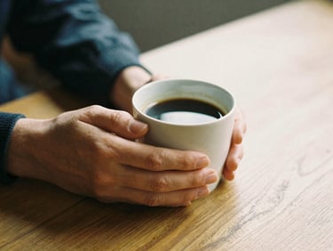 A close-up of hands holding a warm mug of coffee on a wooden table, soft overcast daylight, peaceful atmosphere.