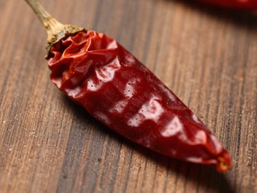 Extreme close-up of a single dried red chili pepper on raw wooden surface, north-facing daylight, deep rust color, texture visible on skin, no other props