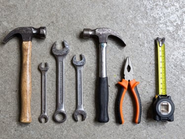 Head-on flat lay of hand tools — hammers, wrenches, pliers, and tape measures arranged in a row on a concrete floor, bright even warehouse lighting, no shadows obscuring tool markings