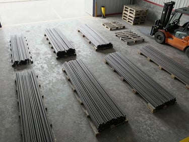 Overhead view of an organized loading bay with steel bar bundles and cement pallets arranged on a concrete floor, bright even daylight from open bay doors, forklift visible at far edge, clean industrial framing showing material density and scale