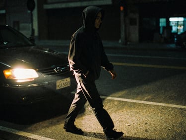 Candid motion portrait of a subject in a dark streetwear hoodie walking briskly under harsh yellow headlights, heavy film grain, dark shadows.