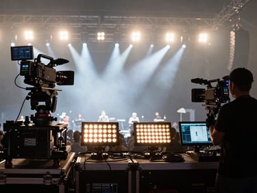 Extreme wide shot of a live music concert broadcast setup, multi-camera rig on a dolly at the far left, another camera operator in silhouette at stage edge right, stage lights cutting through haze in the background, amber tungsten wash on the equipment in the foreground, cables and monitor feeds visible
