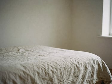 A close-up of a linen-draped bed in an airy room with limewashed walls, soft overcast daylight, desaturated earth tones, 35mm film grain.