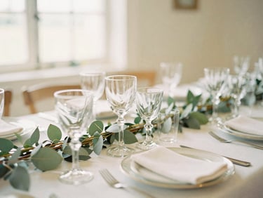 Close-up of an elegant wedding table setting with antique glassware, white linens, and soft sage green foliage, warm diffused window light, 35mm editorial photography