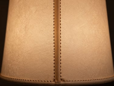 Macro detail of hand-stitched leather seam on a lamp panel, natural tan hide texture in soft north-facing studio light, straight-on material study