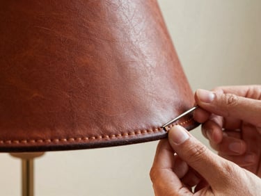 Macro detail of a leather-crafted lamp shade — artisan hands visible at the edge stitching a seam with a curved needle, warm north-facing studio light catching the burnished grain of the hide, stitching thread in sharp focus, background falling to soft blur