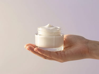 Hands holding a delicate glass jar of botanical cream, soft diffused daylight, warm cream and light purple background
