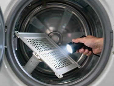 Technician's hand pulling out a dryer lint trap and examining the duct with a flashlight in a laundry room, window light from the side, machine open