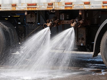 Close-up shot of high-pressure water jets spraying the rusty undercarriage of a semi-truck, heavy water mist, dramatic industrial lighting.