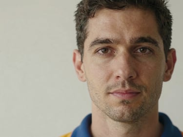 Close-up natural-light portrait of a male plumber in his 30s, direct eye contact, plain light wall behind, honest and confident expression, no studio backdrop