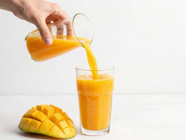 Close-up of hands pouring a thick, vibrant orange mango smoothie into a clean glass, fresh fruit slices nearby, bright daylight