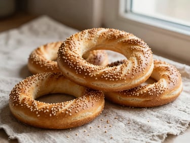Close-up of golden Turkish simit bread rings stacked on a rustic linen cloth, sesame seeds scattering, warm side light.