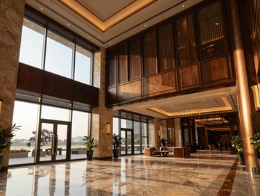 Luxury hotel lobby with high ceilings, polished marble floors, warm bronze accents, and floor-to-ceiling glass windows showing late afternoon light.