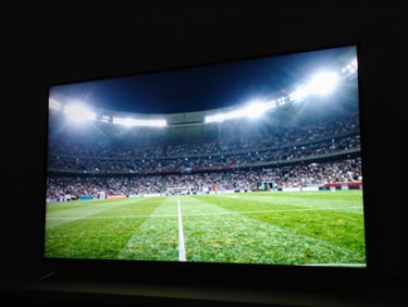 Low-angle high-contrast shot of a glowing smart TV screen in a pitch-black room, displaying a vibrant green football pitch under bright floodlights, dramatic backlighting.
