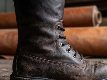 Detail shot of the double-stitched leather seams on a safety boot, showcasing the texture and craftsmanship, against a backdrop of raw leather rolls in an industrial factory, high-contrast lighting, 35mm.