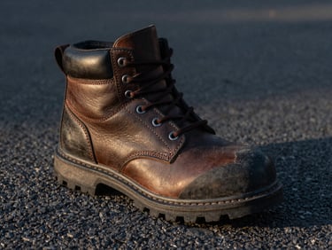 Safety boot on a hot asphalt surface, showing resistance to extreme conditions, with dramatic side-lighting highlighting leather texture and heavy treads, 35mm.